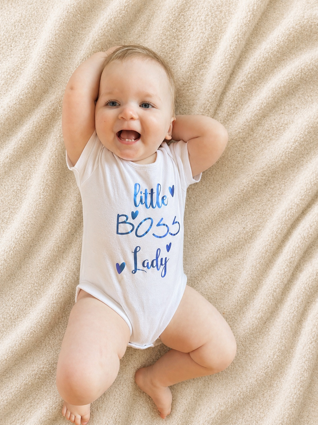 Baby girl lying on cream blanket wearing Little Boss Lady white bodysuit with blue foil print smiling