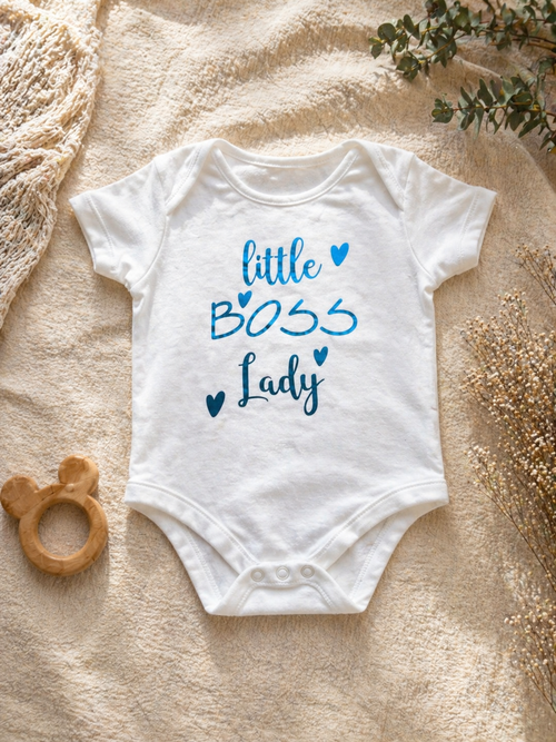White Little Boss Lady baby bodysuit with blue foil heart print laid flat on cream textured background with dried flowers and wooden teether