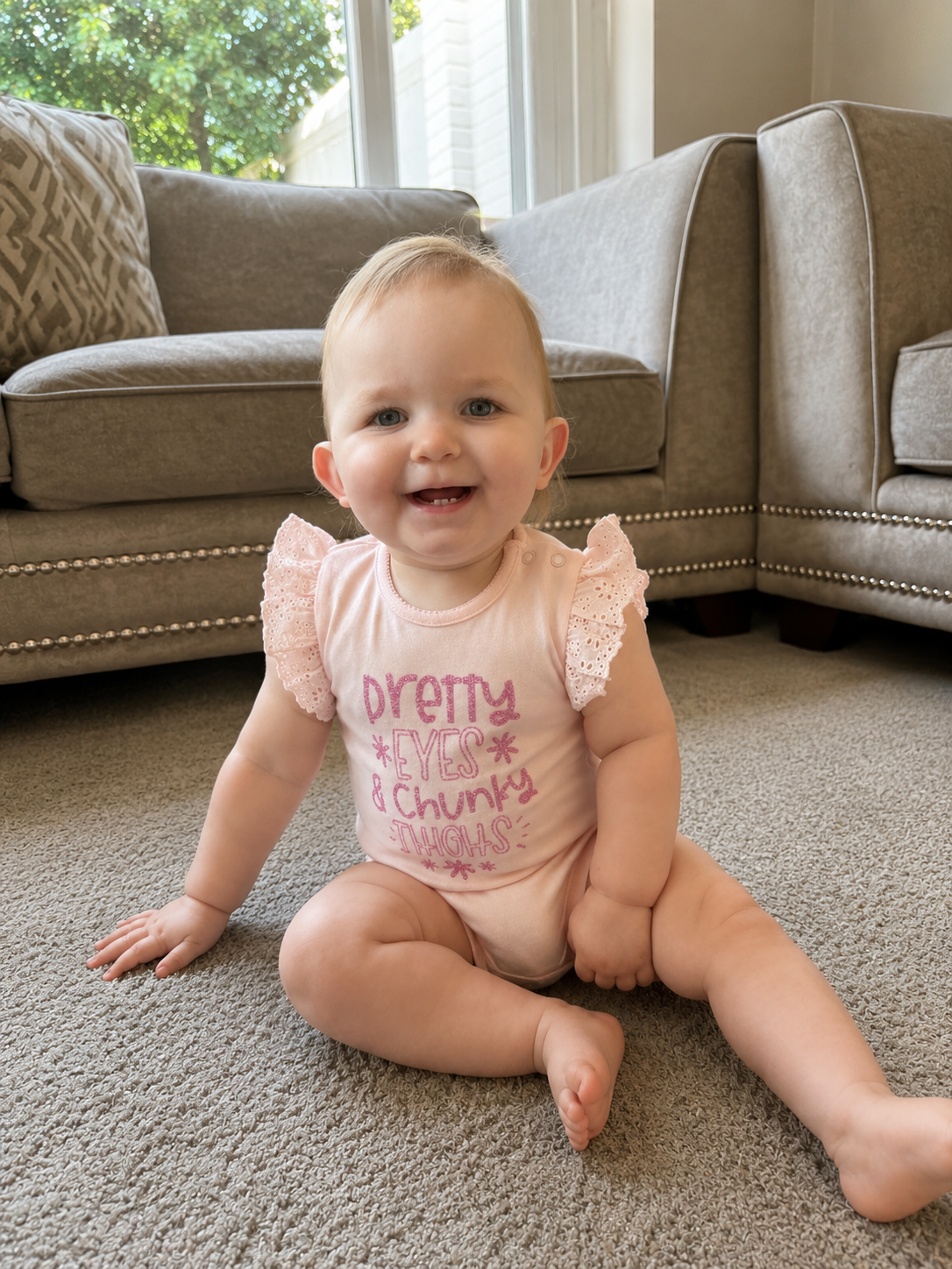 Baby girl sitting on carpet wearing Pretty Eyes and Chunky Thighs pink frill bodysuit smiling