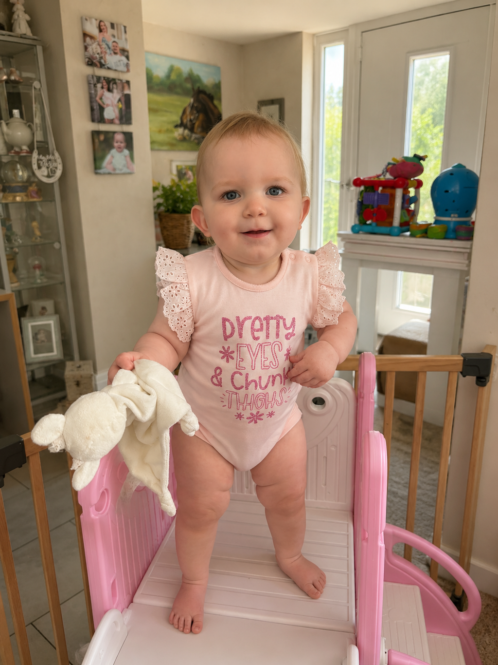 Baby girl standing in pink play area wearing Pretty Eyes and Chunky Thighs pink frill bodysuit